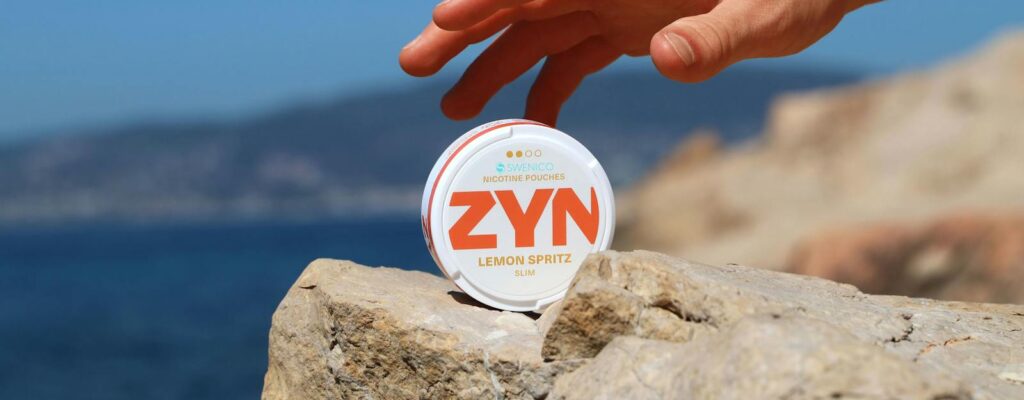 Close-up of a hand reaching for a ZYN nicotine pouch on a rock by the sea.